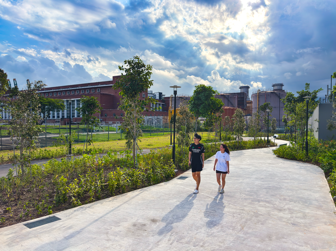 Two people walk on a path in a park with industrial buildings and a dramatic sky.